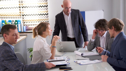 The head of the company conducts a briefing in the meeting room. The man gives instructions to the group. The company's staff sits at a white table