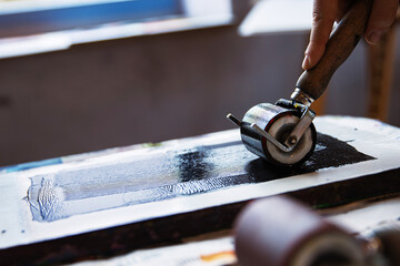 A closeup of an artist hand using a brayer to apply black ink onto a linocut surface in a linocut printmaking process.