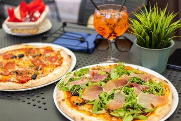 Italian delicious pizza with prosciutto and ham and summer Spritz cocktail in a glass with ice and straws on a table in a restaurant or cafe