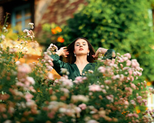 Confident young woman in a green dress flipping her hair back in a blooming garden, surrounded by...