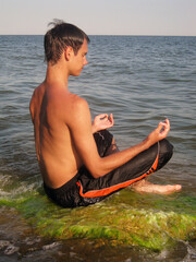 A guy sits in the lotus position on a rock against the backdrop of the sea and meditates