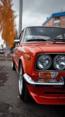 A classic orange car is parked on a city street, showcasing its shiny chrome details. Autumn leaves surround the scene, complementing nearby urban buildings