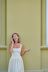Happy elegant young woman talking on mobile phone leaning against a green wall, gesturing with hand