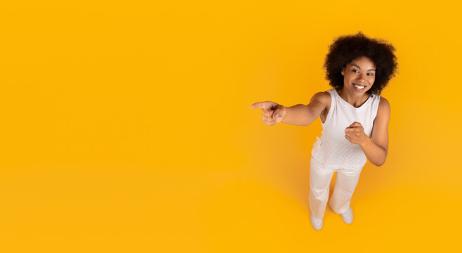 Cheerful young black woman standing and pointing aside with both hands while smiling. Playful high angle shot on yellow background, banner with copy space