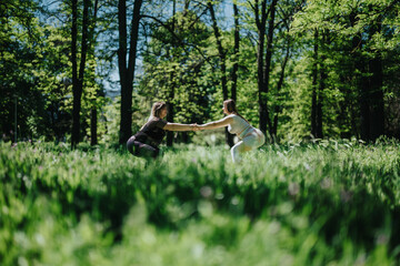 Two women squat opposite each other in a lush forest, enjoying fitness and wellness outdoors,...