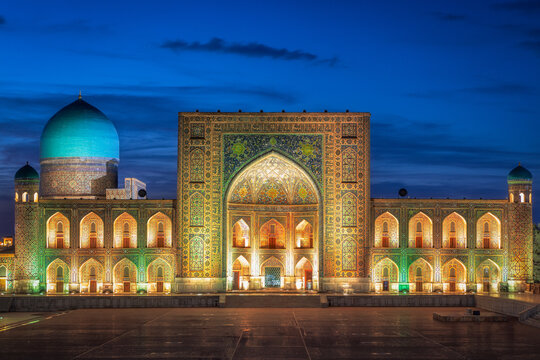 Samarkand, Uzbekistan - 05 July 2024: View of the illuminated Bibi Khanum Mosque against the deep blue twilight sky, showcasing intricate mosaic work and a grand turquoise dome.