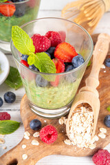 Overnight matcha oatmeal, sweet tasty oats with yogurt or non-dairy creamer, with fresh strawberries, blueberries, raspberries and mint, with ingredients on white wooden kitchen table