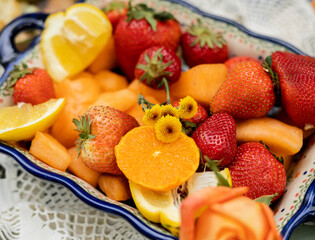 Fresh strawberries, orange slices, and melon in ceramic dish with lemon wedges and flowers
