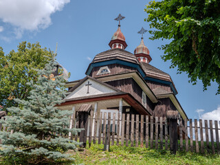 Front view of wooden church in Nizny Komarnik, Slovakia. Peaceful rural scene with trees, fence and cross, showing traditional Carpathian architecture in summer light.
