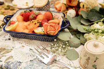 ceramic dish filled with strawberries, melon cubes, lemon slices, and decorated with orange roses and flowers on a lace tablecloth for an outdoor garden party