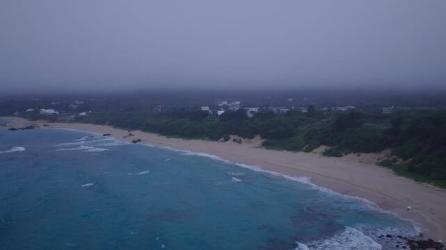 Flying over blue water and sandy beach with coastal houses and fog at dawn