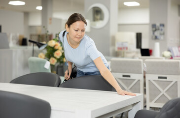 Fototapeta premium Curious young woman choosing functional dining table for her home in furniture shop, carefully examining display samples..