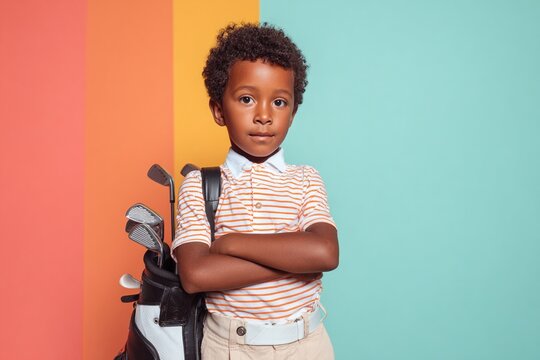 Young african american boy with curly hair, wearing striped shirt and beige pants, confidently poses with golf clubs and bag against colorful background, showcasing passion for golf
