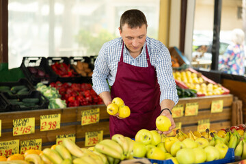 Man seller of garden stuff department store is replenishing showcase with fruits, puts apples on showcase. Male employee was distracted for minute and stands smiling with ripe apple in hands.