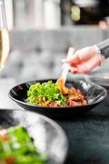 Close-up of a hand using chopsticks to pick up food from a black bowl filled with colorful dish and cilantro, with blurred background, capturing a vibrant dining moment.