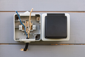 Outdoor electrical outlet with open cover and exposed wires mounted on gray wooden wall...