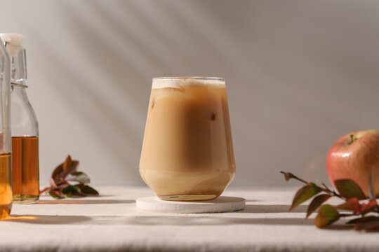 Chilled apple latte in a modern glass stands on a sunlit table, surrounded by golden syrup bottles and autumn branches, evoking cozy fall vibes.