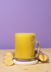 Bright glass mug with golden turmeric latte on wooden surface, fresh sliced ginger and whole root nearby, vivid purple background for health and wellness concept.