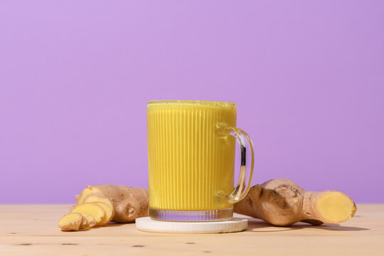Glass mug filled with vibrant golden turmeric latte on wooden table, fresh ginger roots nearby, bright purple background creates bold contrast for healthy drink.