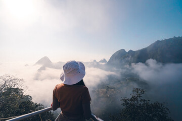 A woman standing on a mountain with sunrise in a soft tone. She turned around and looked at the beautiful view. There is a forest below on a foggy day.  Idea for nature wallpaper with copy space.