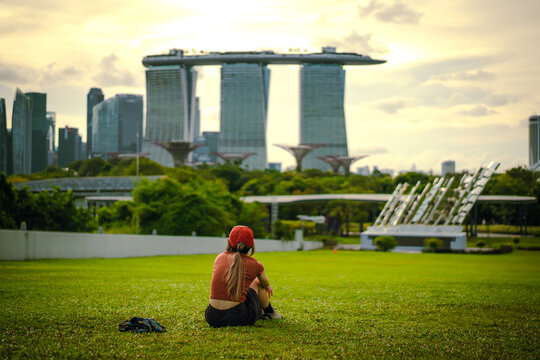 Front view of a woman sitting on the lush green lawn, looking at the view of Marina Bay Sands in Singapore. There are trees in the background and a park with no people in the evening on a cloudy day.