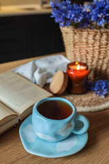 Blue ceramic cup with white cloud pattern on saucer, standing on wooden kitchen table. Nearby an open book, candle in amber jar, and a basket filled with blue cornflowers add cozy ambiance