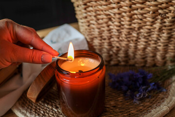 Female hand lighting a candle with a matchstick in a cozy kitchen setting. The candle is in an amber glass jar near a woven basket filled with blue cornflowers, evoking a warm, intimate mood
