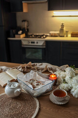 Fresh cinnamon rolls with icing in a glass dish on a wooden kitchen table. Nearby are a lit candle, open book, cup of tea, teapot, and white peonies. Cozy home baking atmosphere