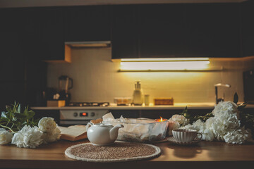Cozy kitchen scene with a white teapot, a cup of tea, cinnamon rolls in a glass dish, blooming white peonies, a burning candle, and an open book on a wooden table