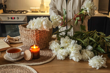 Person arranging fresh white peonies into a woven basket on a wooden kitchen table. Nearby are a...