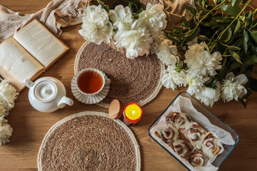 Flat lay of a cozy breakfast table with cinnamon rolls, white peonies, a teacup, teapot, an open book, and a glowing candle, styled in soft natural tones and textures
