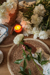 Close-up of a child's hand holding a white peony flower on a rustic placemat, with candlelight, woven textures, and a bunch of fresh peonies in the background, creating a cozy floral mood