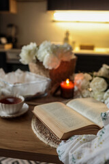 Open book on a wooden table beside a cup of tea, candle, and peonies in a cozy kitchen setting. Soft lighting and woven textures create a warm, peaceful reading atmosphere