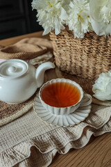 Close-up of a ceramic cup with tea and teapot on textured fabric and wooden table. White peonies in a wicker basket add a rustic and cozy touch to this calm kitchen scene