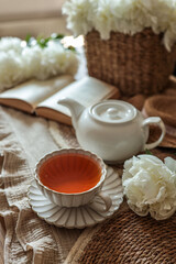 Cozy tea setting with a ceramic cup filled with tea, a white teapot, blooming white peonies, and an open book on a soft fabric runner. Warm, rustic atmosphere in natural light