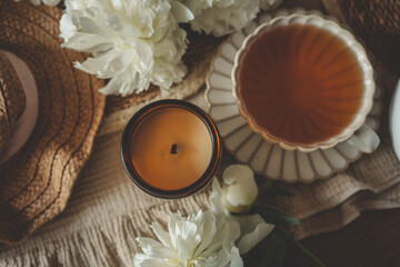 Aromatherapy candle in amber jar, cup of tea and white peonies in cozy summer flat lay with straw...