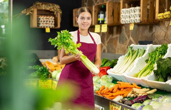 Young woman seller of garden stuff store is replenishing showcase with vegetables, puts greenery celery on showcase. Girl worker was distracted for minute and stand smiling with celery in hands.
