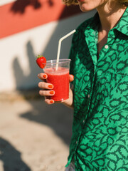 A vibrant red smoothie with a strawberry garnish held by a person in a green patterned shirt, perfect for a sunny day refreshment.