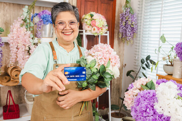 Senior woman, florist shop, small business owner holds credit card with confidence, bouquet in...
