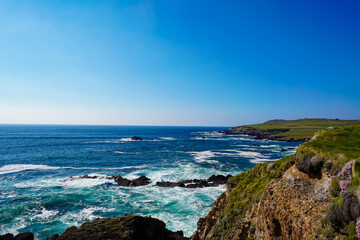 Sunny coastal scene with rocky cliffs, turquoise waves, and green meadows overlooking the Atlantic Ocean.