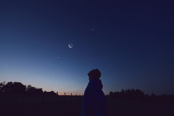 Silhouette of a man observing Moon, Milky Way stars, planets and other celestial objects from a dark countryside location.