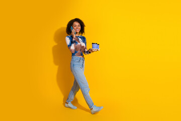 Young stylish woman with curly hair walking while holding coffee and talking on mobile phone against vibrant yellow background