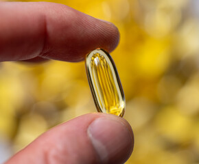 Single transparent yellow fish oil capsule held between fingers with blurred golden capsules background. Dietary supplement and vitamin concept. Macro shot photography. 