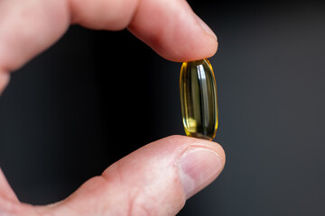 Yellow softgel capsule held between fingers on dark background. Close-up studio shot of transparent dietary supplement. Healthcare and pharmaceutical concept for design and banner