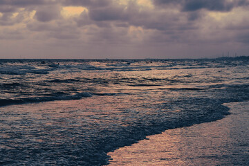 Obraz premium A serene sunset over a wavy ocean, reflecting purple and orange hues from the cloudy sky onto the water, with a distant coastline visible.