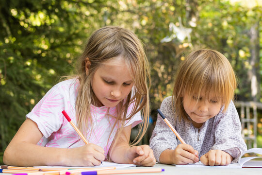 Two young sisters drawing with colorful fineliners in notebooks at an outdoor table - Powered by Adobe