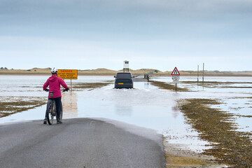 woman with bicycle watching a car, crossng the flooded Causeway to Holy Island , Northumbri,...