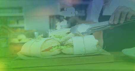 cook in apron arranging two wrapped baguette sandwiches with tongs on board at cafe kitchen counter