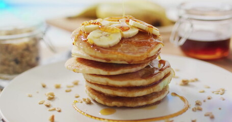 Pancake stack sitting on kitchen table with banana slices and syrup, with granola jar