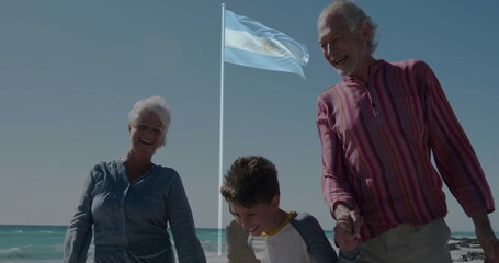 Walking seniors and grandson holding hands along sandy beach, striped flagpole and ocean waves